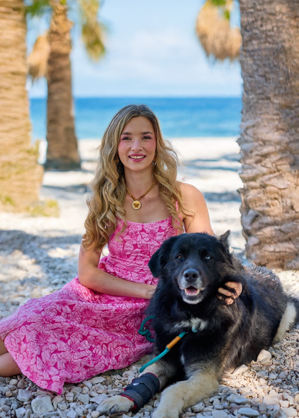 Laura im pinken Kleid am Strand unter Palmen, neben ihr ihr Hund Sky